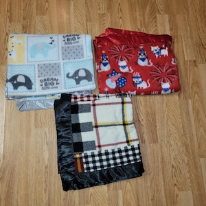 May include: Three fleece blankets with different patterns. The first blanket is blue and gray with a white elephant and the text "Dream Big Little One". The second blanket is red with a white cat and fireworks. The third blanket is black and white plaid with a black satin border.