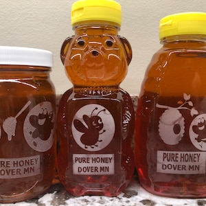May include: Three glass jars of honey with yellow lids. The jars are decorated with a white label that says "PURE HONEY DOVER MN" and a cartoon bee design. The honey is a light amber color.