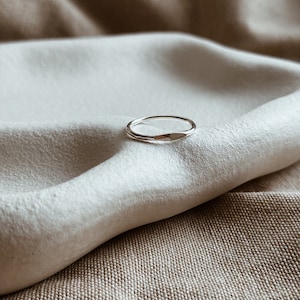 May include: A simple silver band ring with a slightly rounded, flat top. The ring is resting on a white and brown fabric background.