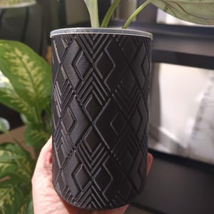 Könnte beinhalten: Ein schwarzer, zylindrischer Pflanzentopf mit einem geometrischen Rautenmuster. Er enthält eine grüne Pflanze mit großen, geäderten Blättern. Der Topf wird von einer Person in der Hand gehalten.