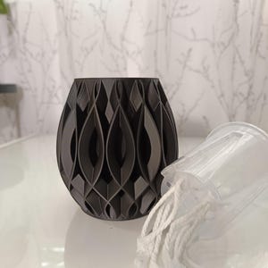 May include: A dark gray, decorative vase with a geometric, openwork design. The vase is cylindrical with a rounded base. A clear plastic container with white rope is next to the vase. The background is a white curtain.