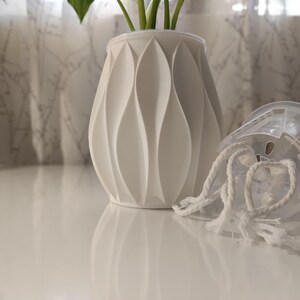May include: A white, decorative vase with a textured, geometric design. A clear plastic pot with white rope handles sits next to the vase. The vase is holding green plants. The objects are on a white surface.