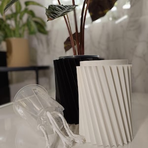 May include: A black and white geometric planter with a clear plastic inner pot and a plant. The planters have a ridged design. The clear pot has white rope-like wicks. The plant has dark green and brown leaves.