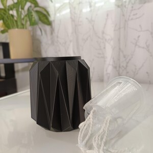 May include: A black geometric planter with a clear plastic pot and white rope. The planter has a faceted design. A green plant in a beige pot is in the background.
