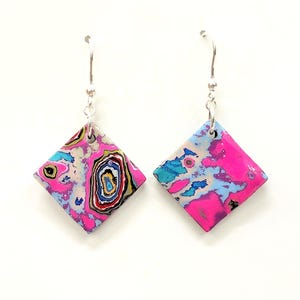 Könnte beinhalten: Ein Paar lebendige, abstrakte Ohrringe in Diamantform. Die Ohrringe weisen eine Mischung aus rosa, blau und beige Farben mit einem silbernen Haken auf. Das Design hat ein einzigartiges, wirbelndes Muster und verleiht jedem Outfit Farbe.
