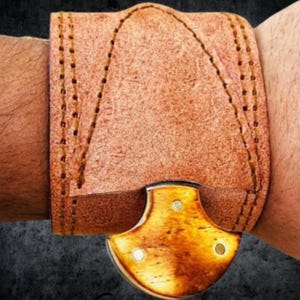 Puede incluir: Primer plano de una pulsera de cuero marrón con un cuchillo decorativo. La pulsera está cosida con un hilo en contraste. El cuchillo tiene un mango dorado con tres remaches blancos.