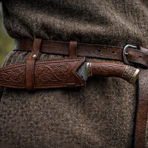 May include: A brown leather sheath with an ornate Celtic knot design holds a knife with a wooden handle and a metal blade. The sheath is attached to a brown leather belt with a silver buckle. The person is wearing a brown tunic.