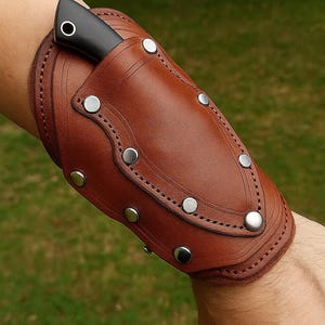 Puede incluir: Una funda de cuchillo de cuero marrón con un cuchillo negro parcialmente insertado. La funda está asegurada con remaches plateados y detalles cosidos. El diseño está curvado para adaptarse al brazo.