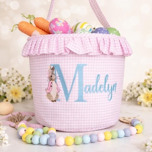 May include: Pink gingham Easter basket with a ruffle trim, filled with decorative eggs and carrot-shaped items. The basket features a blue embroidered initial "M" and the name "Madelyn", along with a bunny illustration. A pastel bead garland is in the foreground.