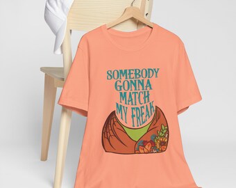 May include: Peach-colored t-shirt with the text "SOMEBODY GONNA MATCH MY FREAK" in teal. The graphic features a stylized design with wings and flowers. A white baseball cap rests on a wooden chair behind the shirt.