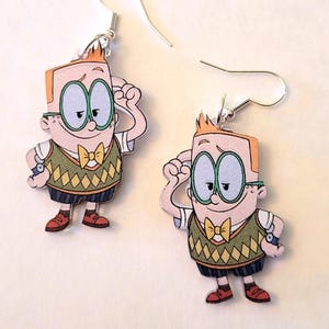Może przedstawiać: Para kolczyków z postacią z kreskówki z pomarańczowymi włosami, zielonymi okularami i zieloną kamizelką. Kolczyki mają srebrne haczyki i są umieszczone na białym tle.
