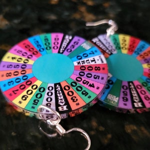 May include: A pair of earrings shaped like a colorful wheel of fortune with numbers and the word "BANKRUPT" on it. The earrings are silver and have a hook closure.