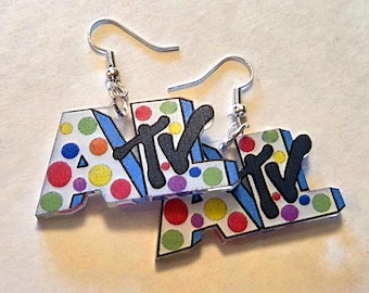 May include: A pair of drop earrings featuring the letters MTV in a stylised font. The letters are white with colourful polka dots in red, yellow, green, blue, and purple. A large black letter M overlaps the white letters, and silver hooks are attached.