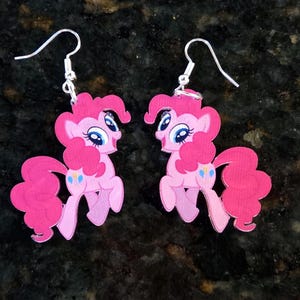 Könnte beinhalten: Ein Paar Ohrhänger mit einem rosa Pony-Charakter, einer lockigen rosa Mähne und einem Schweif, großen blauen Augen und drei kleinen blauen und gelben Ballonsymbolen auf der Flanke. Die Ohrringe sind an silberfarbenen Metallhaken befestigt.