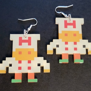 May include: A pair of pixelated robot earrings with a white body, orange legs, and green shoes. The robot has a yellow head with a red "H" on the top.