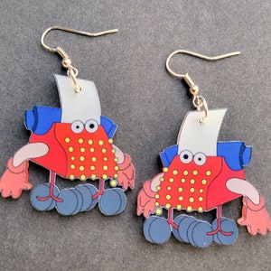 Puede incluir: Un par de pendientes rojos, azules y blancos con un personaje de máquina de escribir de dibujos animados con puntos amarillos. Los pendientes tienen ganchos dorados.
