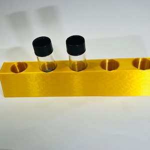Puede incluir: Un soporte rectangular de color dorado con cuatro aberturas circulares. Dos pequeños frascos de vidrio con tapas negras están en el soporte. El soporte está hecho de un material texturizado y se encuentra sobre una superficie blanca.