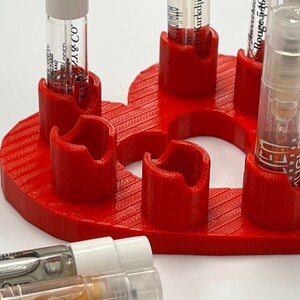May include: Red heart-shaped holder with six slots for perfume sample vials. The holder is made of 3D printed plastic and has a textured surface.