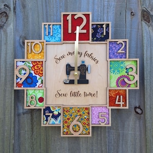 Może przedstawiać: Drewniany zegar ścienny z motywem maszyny do szycia. Tarcza zegara zawiera kolorowe kwadraty z tkaniny z drewnianymi cyframi. Tekst brzmi "Sew many fabrics, sew little time!" Złote wskazówki wskazują godzinę.