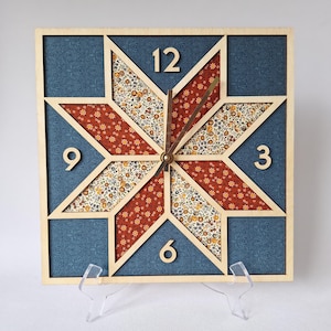 May include: Square wooden wall clock with a starburst design. The clock face features a blue floral patterned background, with a star-shaped design in red and floral fabric. The clock hands are brown, and the numbers 3, 6, 9, and 12 are visible.