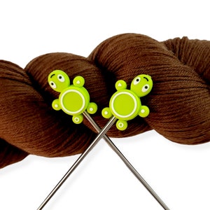 May include: Two silver knitting needles with green turtle-shaped stitch markers. The needles are crossed and the yarn is brown.