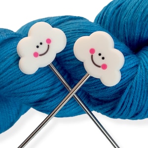May include: Two knitting needle toppers with white cloud designs, each featuring a smiling face with pink cheeks and black eyes. The needles are crossed in front of a ball of blue yarn, creating a cheerful and whimsical craft theme.
