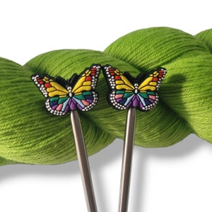 May include: Two butterfly-shaped stitch markers with colorful wings and silver metal stems. The markers are in front of a large skein of bright green yarn, creating a vibrant contrast. The butterfly wings feature yellow, orange, red, purple, and blue details.