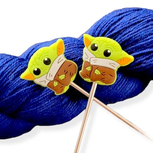 May include: Two knitting needles with green and brown character shaped ends. The needles are resting on a ball of blue yarn.