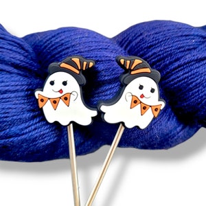 May include: Two white ghost-shaped stitch markers with orange and black hats and orange and black bunting. The markers are on a ball of blue yarn.