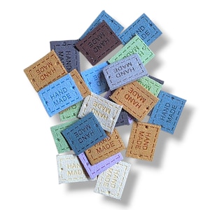 May include: A collection of small, square fabric tags in various colors, including blue, brown, green, and purple. Each tag has the words "HAND MADE" embroidered on it, suggesting they are for handmade crafts.