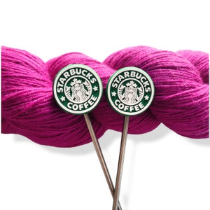 May include: Two knitting needle stitch markers with the Starbucks Coffee logo, set against a vibrant magenta yarn ball. The markers have a green and white logo design and silver needles.