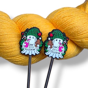 May include: Two decorative Christmas-themed cocktail stirrers. Each stirrer features a white ghost wearing a green hat, holding a pink drink and a gingerbread man. The text "DON'T STOP BELIEVIN" is printed on each stirrer.