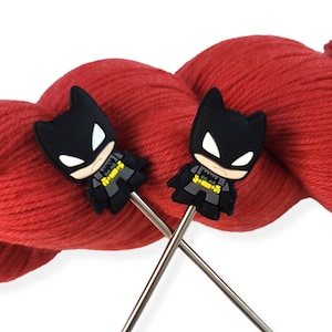 May include: Two silver knitting needles with black Batman character toppers. The needles are crossed and resting on a ball of red yarn.