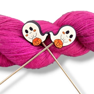 May include: Two silver knitting needles with ghost-shaped stitch markers. The ghosts are white with black eyes and smiles, and each is holding a small orange pumpkin. The needles are resting on a pink ball of yarn.