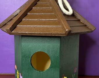 May include: A green wooden birdhouse with a brown roof and a small opening for birds to enter. The birdhouse is decorated with painted flowers in various colors, including yellow, red, purple, and orange.