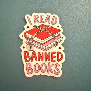 May include: A sticker with the text "I read banned books" in pink and red lettering. The sticker features a stack of books bound together with a chain and padlock.