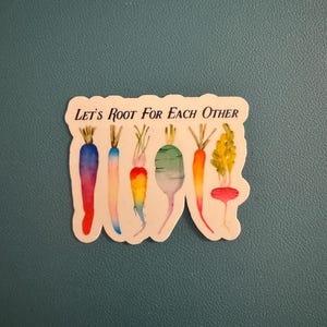 May include: A sticker featuring watercolor illustrations of colorful root vegetables. The text "Let's Root For Each Other" is printed above the vegetables. The vegetables include carrots, radishes, and other root vegetables in shades of red, orange, yellow, blue, and green.