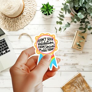 May include: A sticker with the text "DIDN'T SEEK Validation FROM Social Media TODAY" in black and white. The sticker is shaped like a ribbon with a yellow border and blue and rainbow accents. The sticker is held by a hand.