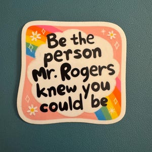 May include: A sticker with a pink border and the text "Be the person Mr. Rogers knew you could be" in black. The sticker has a rainbow, flowers, and diamonds design.