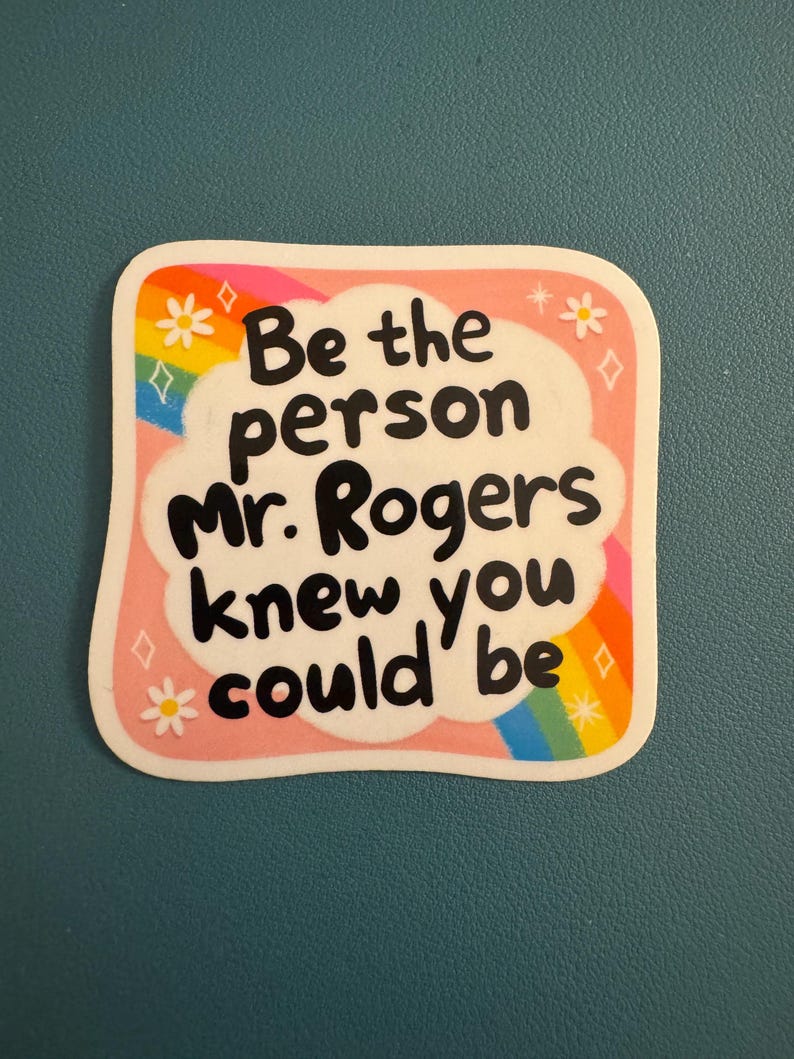 May include: A sticker with a pink border and the text "Be the person Mr. Rogers knew you could be" in black. The sticker has a rainbow, flowers, and diamonds design.