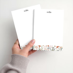 May include: Two white notepads with the word "notes" in black script at the top. The bottom of each notepad features a floral design with orange, green, and brown flowers and leaves. The notepads are held by a hand.