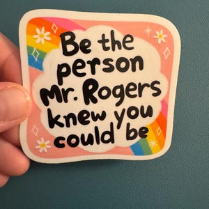 May include: A square sticker with rounded corners. The sticker has a pink border with white cloud-shaped text that reads "Be the person Mr. Rogers knew you could be" in black. Rainbow and floral accents decorate the border.