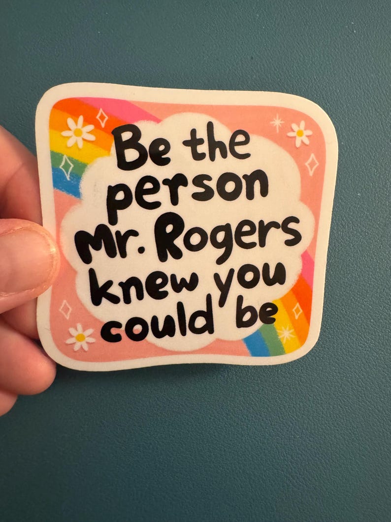 May include: A square sticker with rounded corners. The sticker has a pink border with white cloud-shaped text that reads "Be the person Mr. Rogers knew you could be" in black. Rainbow and floral accents decorate the border.