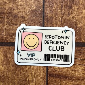 May include: A white sticker with a pink and yellow smiley face, the text "SEROTONIN DEFICIENCY CLUB", the text "VIP MEMBERS ONLY", and a barcode with the number "67938001".