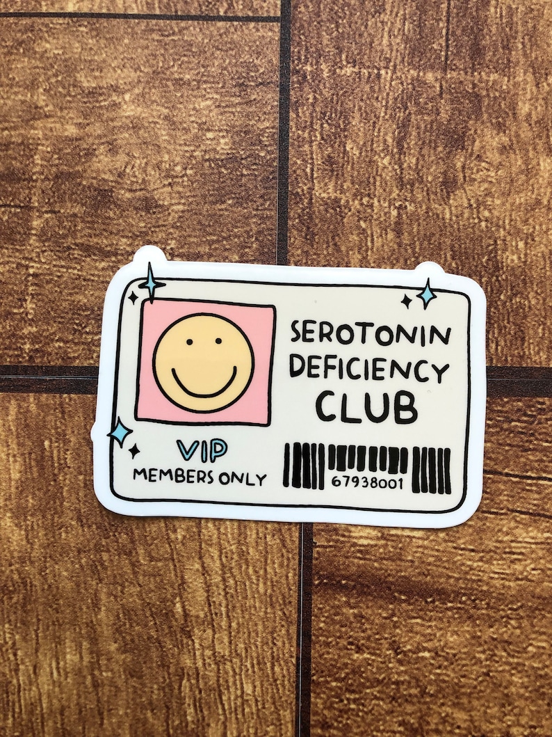 May include: A white sticker with a pink and yellow smiley face, the text "SEROTONIN DEFICIENCY CLUB", the text "VIP MEMBERS ONLY", and a barcode with the number "67938001".