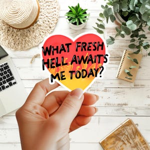 May include: A heart-shaped sticker with a gradient background transitioning from red at the top to yellow at the bottom, featuring orange flame illustrations and the black text: WHAT FRESH HELL AWAITS ME TODAY? The sticker has a white border.