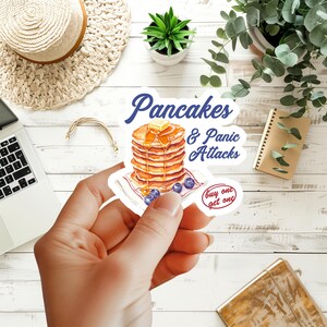 May include: A die-cut sticker featuring a stack of golden-brown pancakes topped with a pat of butter and syrup, surrounded by blueberries. The text reads "Pancakes & Panic Attacks" in blue script and "buy one get one" in a red oval.