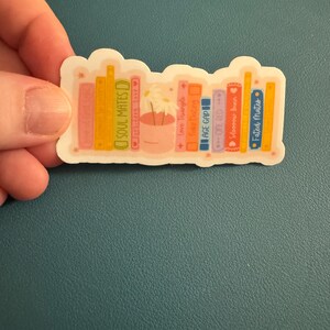 May include: A sticker featuring a bookshelf design with various book titles, including "SOUL MATES," "Love Triangle," and "Fated Mates." The books are in shades of pink, orange, and blue, with a small pot of flowers in the center.