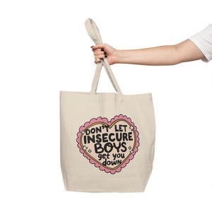 Könnte beinhalten: Eine beigefarbene Tragetasche aus Segeltuch mit einer zentralen Grafik in Form eines rosa gewellten Herzens. Im Inneren des Herzens steht der schwarze Text: DON'T LET INSECURE BOYS get you down. Das Design enthält kleine weiße Sterne und zwei Tragegriffe.