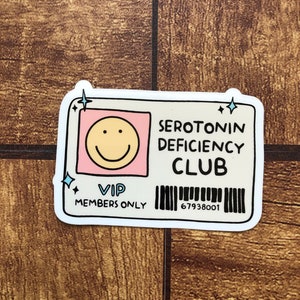 May include: A white sticker with a cartoon smiley face, the text "SEROTONIN DEFICIENCY CLUB", "VIP MEMBERS ONLY", and a barcode with the number "67938001".